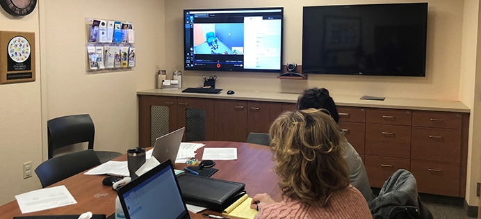 Two people sitting at a conference room table with a computer and note pad watching one of the two TVs on the wall