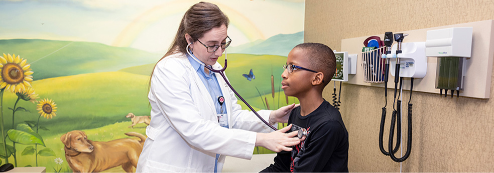 A woman doctor with long blonde hair and glasses in a white lab coat listening a little boy’s heart with a stethoscope in a pediatric exam room that has a mural in the background. The little boy has glasses, black hair that’s cut short, and is wearing a black shirt.