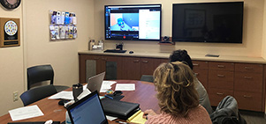 Two people sitting at a conference room table with a computer and note pad watching one of the two TVs on the wall